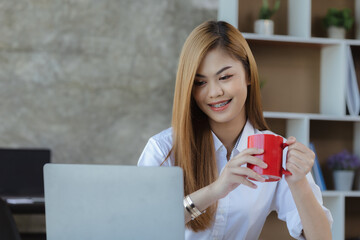 Asian female worker in a corporate office office holding a red coffee mug, she is an employee of a startup finance and tax department. Concept of corporate finance and tax management.