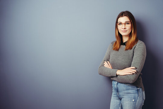 Young woman with glasses and folded arms looks at camera, blue wall with copy space