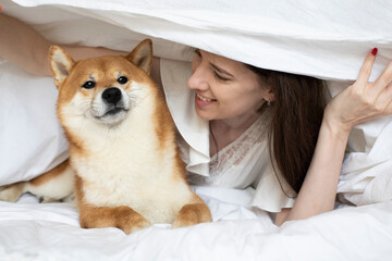 Young funny girl woman playing with her shiba inu set dog in bed in a hotel. Family time friends