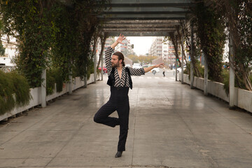 Young man with beard and ponytail, wearing black shirt with white polka dots and black pants and jacket, dancing flamenco in the middle of the city. Concept art, dance, culture, tradition.