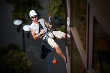 Industrial mountaineering worker in protective helmet showing approval gesture thumbs up while hanging on climbing rope outside building. Man in sunglasses using safety lifting equipment outdoors.