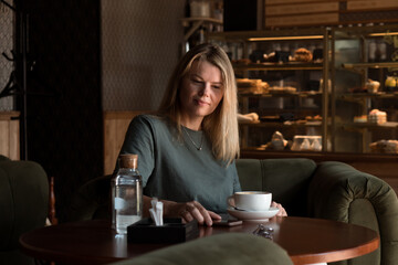 Beautiful blonde woman sits alone in coffeehouse with cup of  coffee and using phone