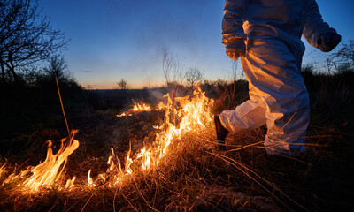 Fireman ecologist fighting fire in field at night. Cropped view of man in protective radiation suit...