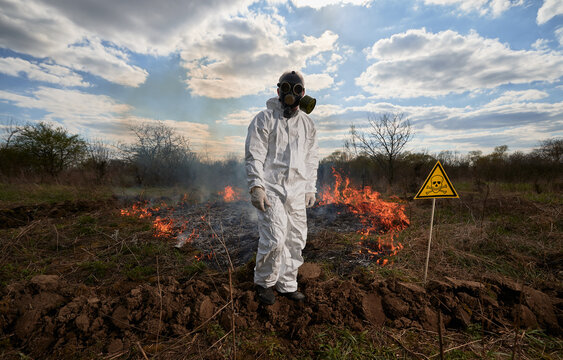 Fireman ecologist working in field with wildfire. Man in gas mask near burning grass with smoke and yellow triangle with skull and crossbones warning sign. Natural disaster concept.