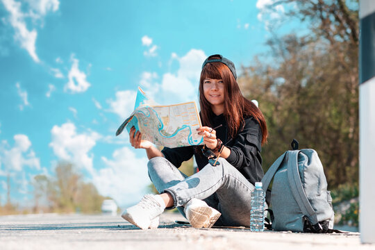 Hitchhiking. A happy hipster girl in cap sitting along the road and holding a paper map. Copy space. Local travel concept