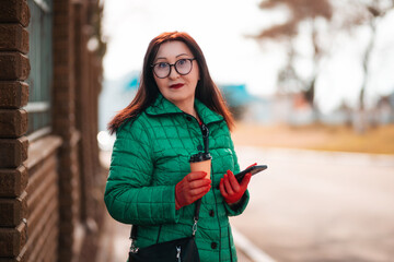 Portrait of a senior beautiful Caucasian woman in glasses and a green jacket holding a cup of coffee and cellphone. Active lifestyle of the elderly