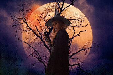 Halloween witch holding magic wand standing over dead tree, full moon and spooky cloudy sky,...