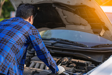 An Asian man looks under the hood of a broken car and checks the oil level on the side of the road.