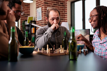 Pensive man sitting at table in living room with multiethnic friends while playing chess. Strained person enjoying strategy boardgames while thinking about chessboard next move.