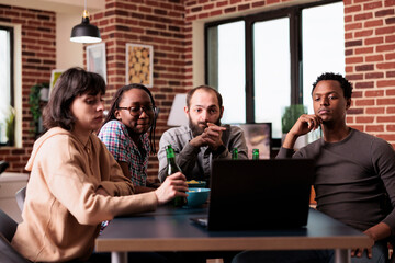 Multiracial group of people sitting at home while watching suspense movies on laptop. Diverse people at table in living room watching online video content together while having snacks and beverages.