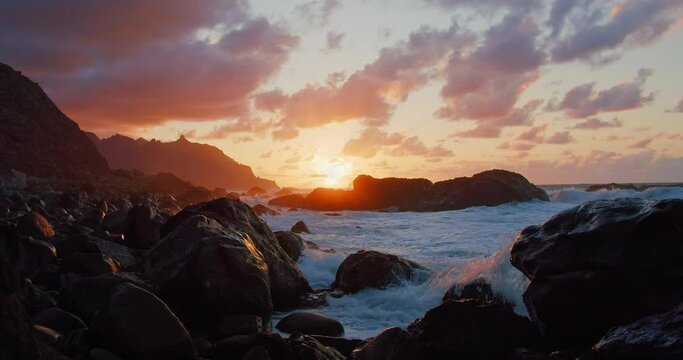 Foaming white sea water sprays over the rugged black rock coast in Tenerife at sunset. Cinematic shot of ocean waves crashing into the volcanic shore of Canary island at golden hour.