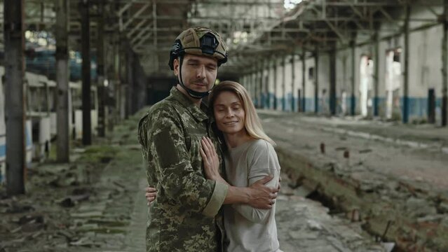 Portrait of male soldier with beloved woman looking at camera while standing in hugs on destroyed factory. Loving couple meeting after long separation.