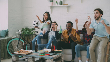 Multi-ethnic group of friends sports fans with Brazilian flags watching football championship on TV together at home indoors and cheering up favourite team - 515574336