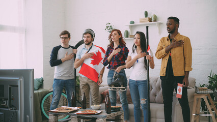 Multi ethnic group of friends sport fans listening and singing national Canadian anthem before watching sports championship on TV together at home indoors - obrazy, fototapety, plakaty