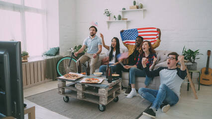 Multi ethnic group of friends with USA national flags watching sport championship on TV together cheering up their favourite team at home indoors