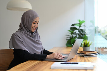 Pleasant Muslim woman using typing business email, searching information on laptop computer