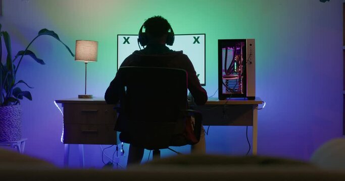 Back view of computer gamer wearing headphones and playing pc games. Teenage boy sitting and using a blank desktop monitor with copy space in a dark room to participate in esports and virtual sports