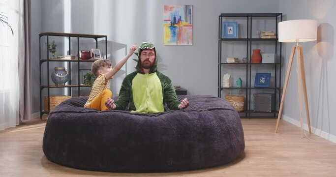 Caucasian handsome man father in green dragon costume sitting on roung soft couch and meditating, then laughing and playing with small cute son. Litle boy having fun with dad at home.