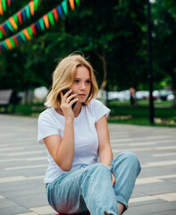 A young beautiful teen girl is sitting on a skateboard on a summer day in the park. A girl is talking on a mobile phone in the open air. Chat with friends.