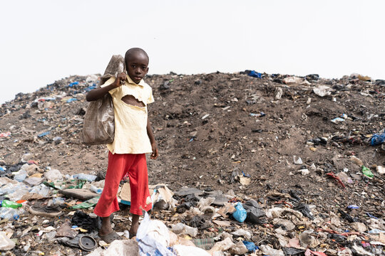 Poor dirty African child working in a landfill with some recycled items in a bag on his shoulders, looking embarrassed at the camera; symbol of extreme poverty in underdeveloped countries