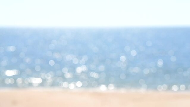 Blurred background. Sea with white sun reflections, yellow sandy beach and blue sky on hot sunny summer day. Sun shines on sea surface water on coast. Beautiful natural background. Seamless Looping