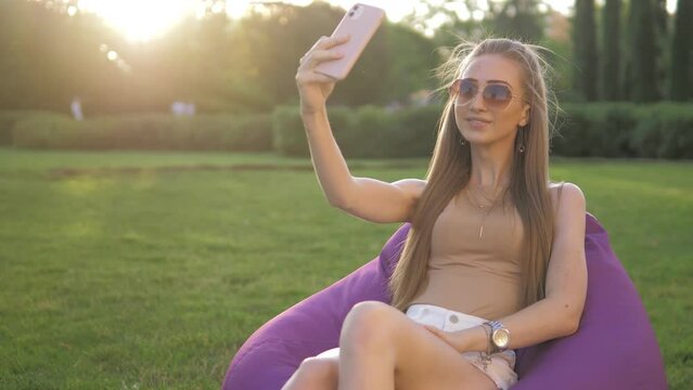 A young beautiful cheerful girl in a well-groomed field at sunset, sitting on a lilac bag chair and looking at her phone. A girl with glasses is resting on a warm bright sunny day. The sun.Green grass