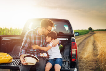 father and son sits on trunk of truck in wheat field. father hugs and kisses his son