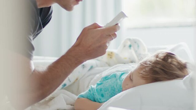 Child care. Father adjusts room's temperature with AC remote control and removes warm blanket from the sleeping kid