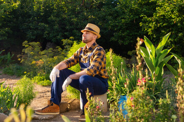 Summertime. A bearded, handsome gardener in uniform and a straw hat sits and relax at his kitchen-garden. In the background there is a backyard and a garden. The concept of gardening and horticulture