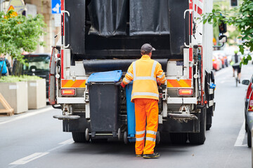 garbage and waste removal services. Worker loading waste bin into truck at city - obrazy, fototapety, plakaty