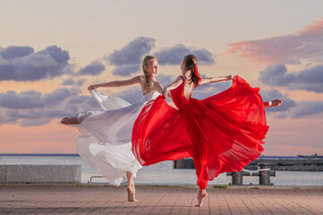 Two ballerinas in a white and red flying skirt and leotard dancing in a duet on the embankment of the ocean or sea against the backdrop of the sunset sky