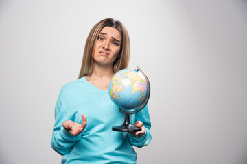 Blonde girl in blue sweatshirt holds a globe and looks uncertain and confused