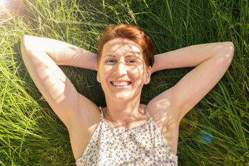 Happy woman lies in the grass in the rays of the setting sun and smiles. Close-up portrait of a woman in summer clothes. The concept of unity with nature, summer vacation. woman smiling