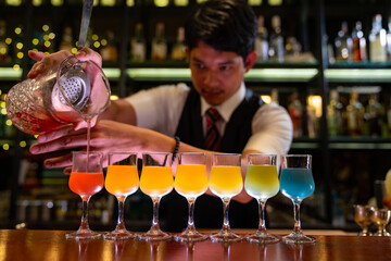 Male bartender pouring mixed colorful cocktail drink from cocktail shaker into shot glass on bar counter at restaurant pub. Mixologist barman making alcoholic drink serving to customer at nightclub