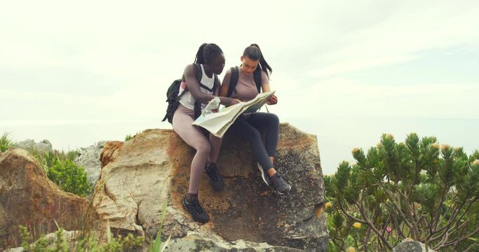 Two women hiking looking at map sitting on a rock against a sky background with copyspace. Friends lost after hike to the top of mountain scenic landscape. Unsure, pointing in different directions