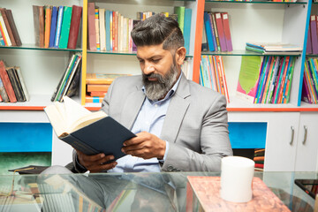 Smart mature indian man wearing suit reading a book while sitting at desk in library with bookshelf...