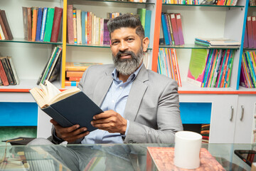 Portrait of smart mature indian man wearing suit reading a book while sitting at desk in library...