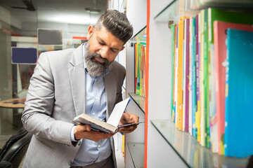 Smart mature indian man wearing suit reading a book at library standing by bookshelf, research,...