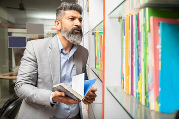 Smart mature indian man wearing suit searching book at library standing by bookshelf, research,...