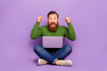 Full length photo of crazy excited man sit floor raise fists celebrate achieve isolated on violet color background