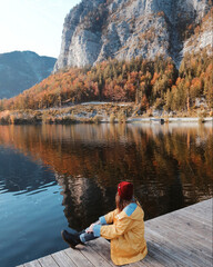 Girl in yellow raincoat sitting near the lake and admiring mountains view. Autumn mountains landscape