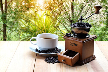 A white hot coffee cup with coffee beans and antique coffee grinders resting on a wooden floor with a tree in the background. Close up. Flare.