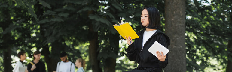 Asian student holding digital tablet and reading book in park, banner.