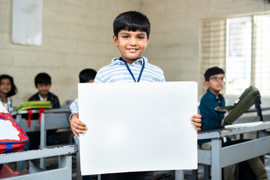 Happy schoolkid showing white empty board by looking camera at classroom in front from to students - concept of promotion, education and advertisement