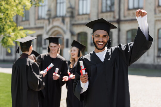 Excited african american bachelor holding diploma near friends in park.
