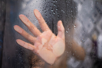 Female hand and unrecognizable silhouette behind wet glass in shower