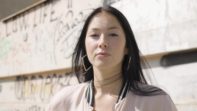 A young beautiful Caucasian woman looks seriously at the camera - closeup - a wall with graffiti in the background