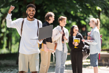 Excited african american student holding laptop near blurred friends in park.