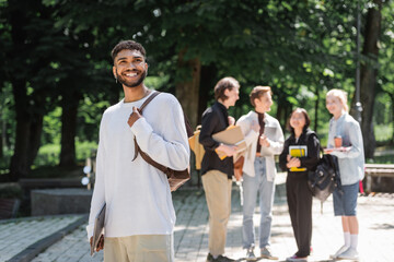 Smiling african american student with laptop standing near blurred friends in summer park.