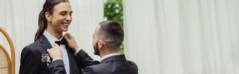 gay man adjusting bow tie on suit of happy groom in formal wear, banner.